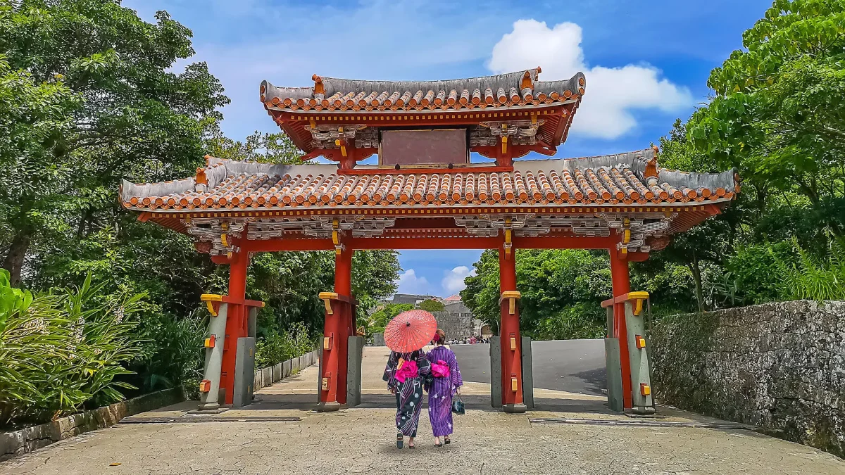 Traditional red Chinese-style gate with tiled roof, flanked by greenery, as two people in kimonos walk beneath, holding a pink umbrella