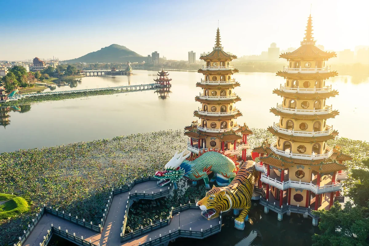 Lakeside temple complex with multi-tier pagodas, a colorful dragon sculpture, and a bridge over a lotus-filled pond at sunrise/sunset fall of light behind.