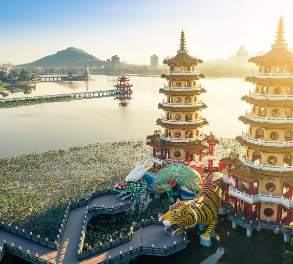 Lakeside temple complex with multi-tier pagodas, a colorful dragon sculpture, and a bridge over a lotus-filled pond at sunrise/sunset fall of light behind.