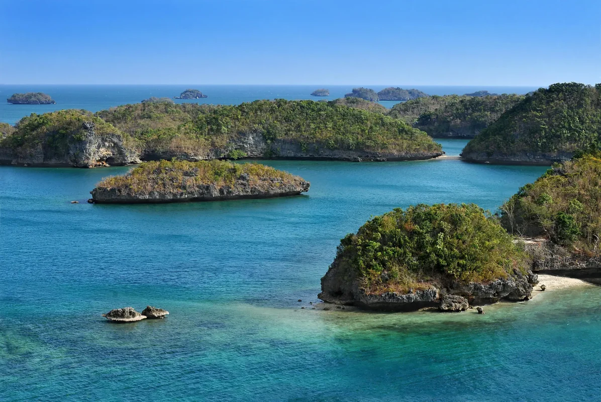 Turquoise coastal scene with rocky green islands and calm water under a clear blue sky, a small sandy cove visible at the right edge.