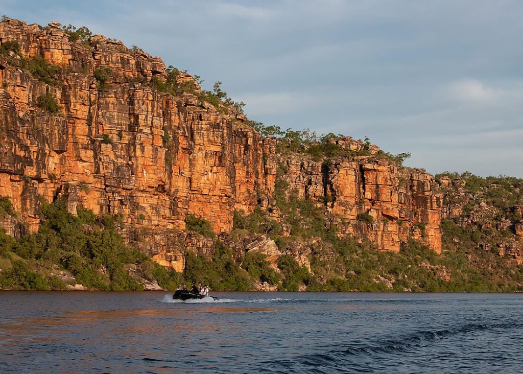 Small boat carrying passengers on calm water beside tall orange sandstone gorge cliffs.