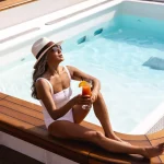 Woman in a white swimsuit and sun hat holds an orange cocktail beside a ship pool.