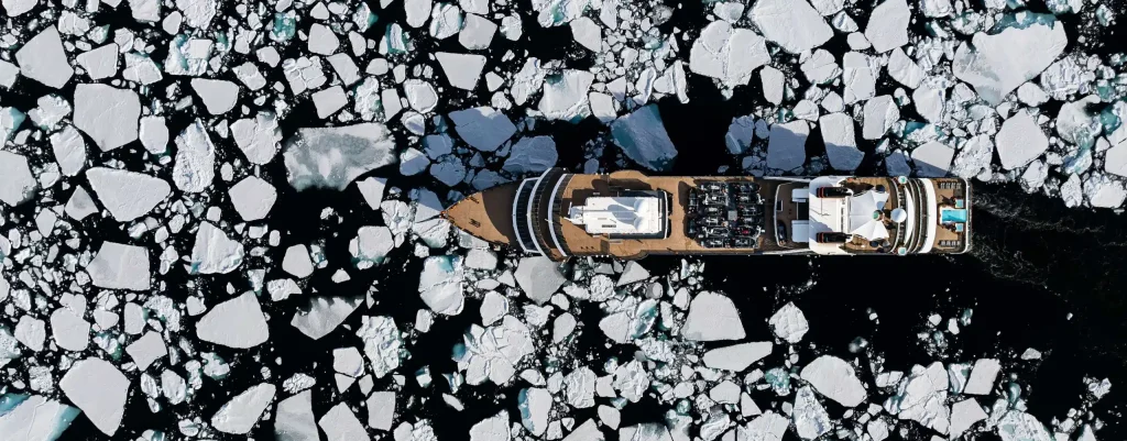 Aerial view of an expedition ship cutting through floating ice chunks in dark water.