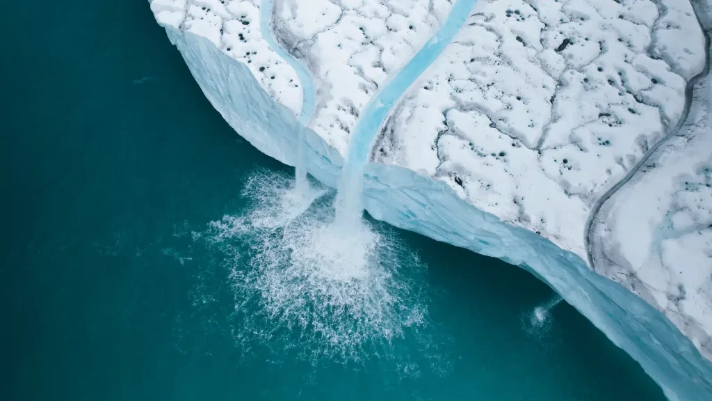 Aerial view of a glacier calving meltwater into teal water beside snowy ice cliffs.