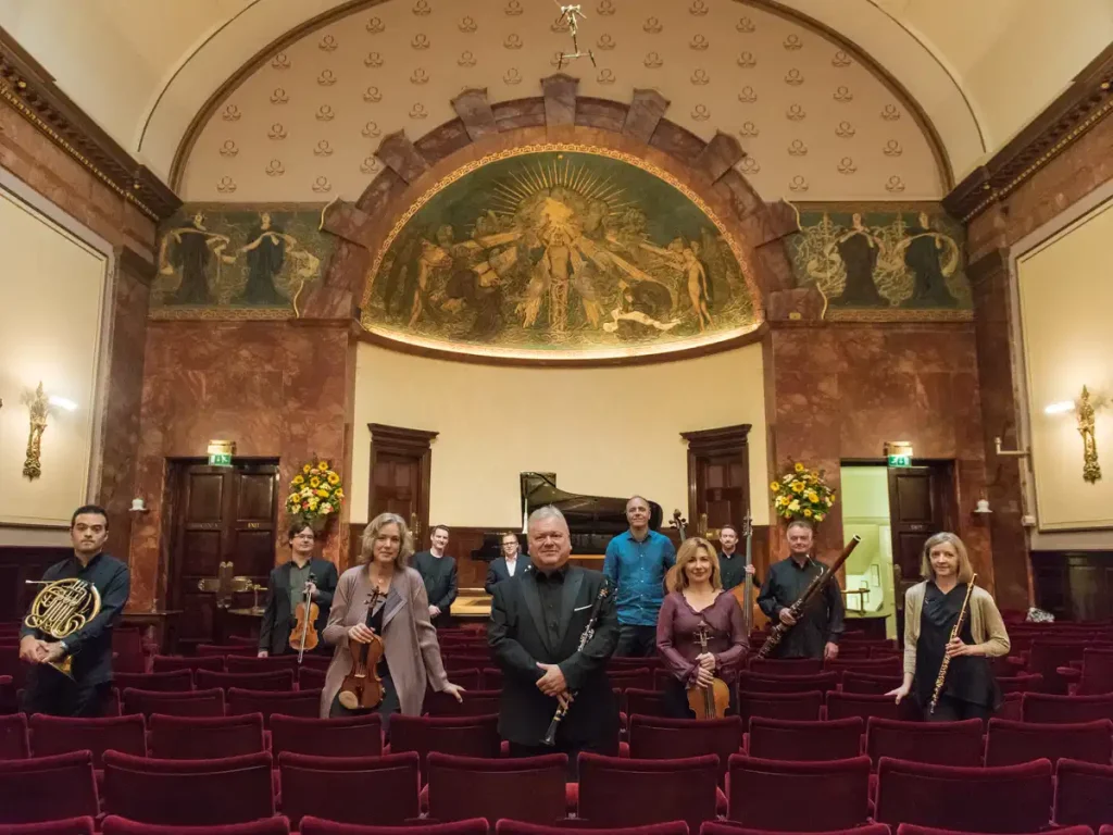 Nine musicians stand in a red-velvet concert hall with a mosaic arch, holding their instruments (violin, cello, flute, clarinet, horn, bassoon).