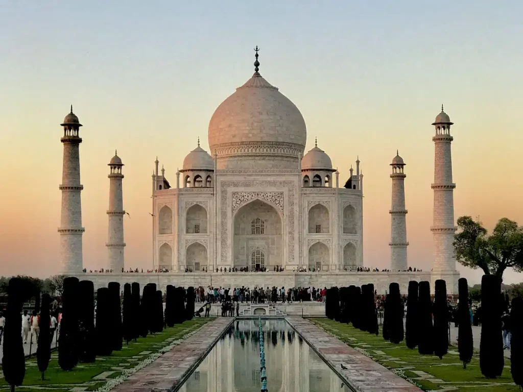 Sunset view of the Taj Mahal, a white marble mausoleum with a central dome and four minarets, mirrored in the front reflecting pool with visitors nearby.