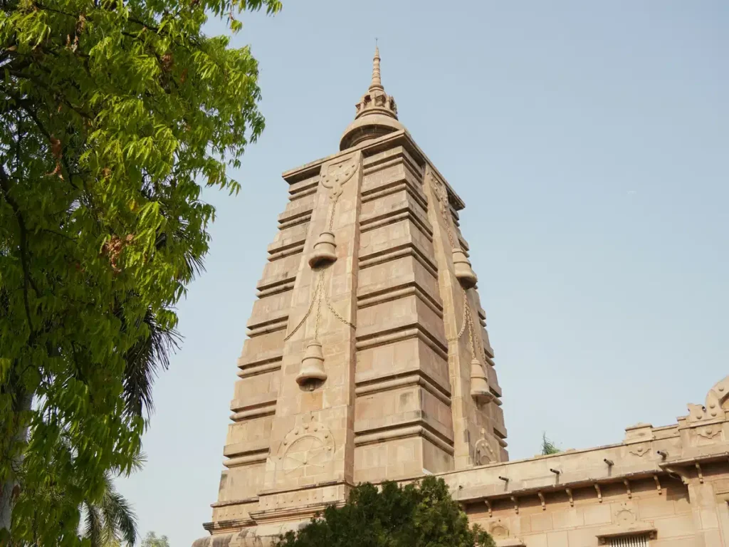 Stone temple tower with tiered architecture rises beside green trees under a clear blue sky, showcasing traditional carvings and a spire at the top.