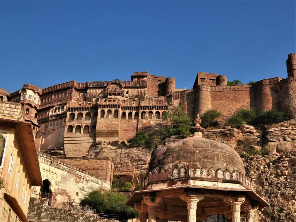 Massive sandstone fortress atop a rocky hill with arches and towers, seen behind a domed pavilion in the foreground under a clear blue sky.