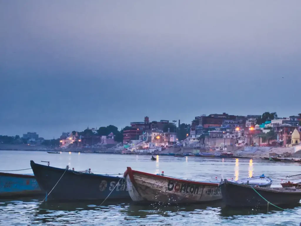 Boats tied near a calm river with a lit town on the far shore at dusk or early evening.