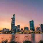 City skyline at dusk with tall skyscrapers rising along a river.