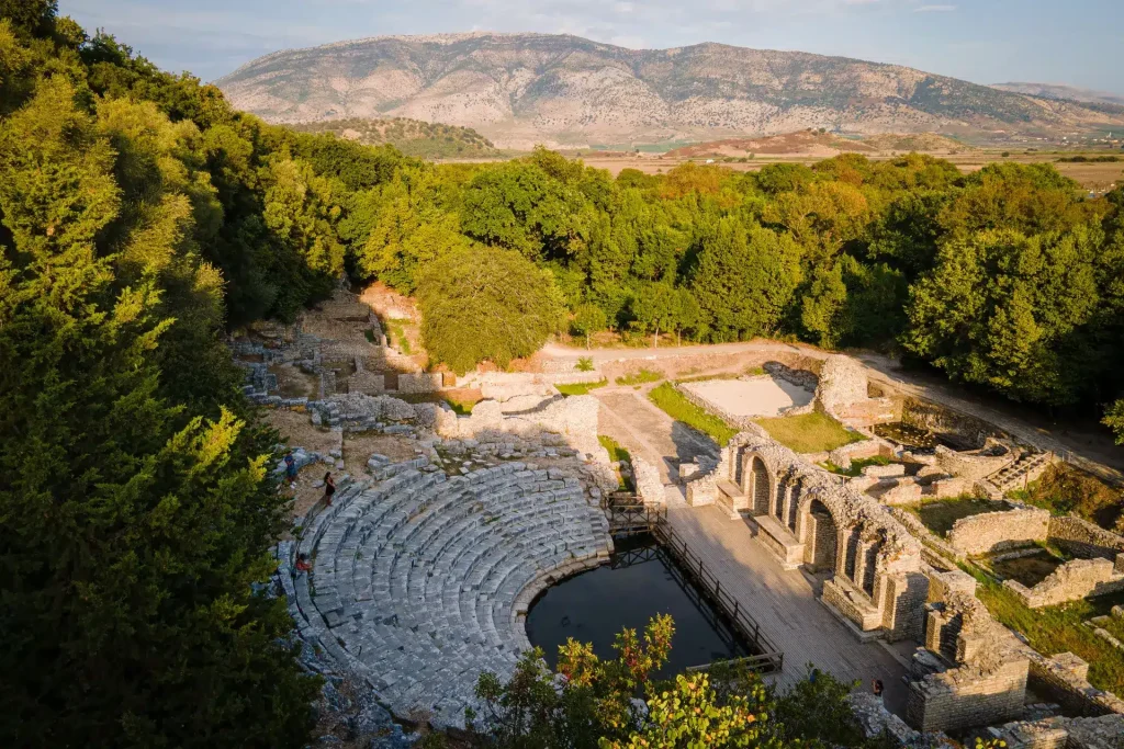 Ancient stone amphitheater surrounded by dense trees with mountains in the distance.