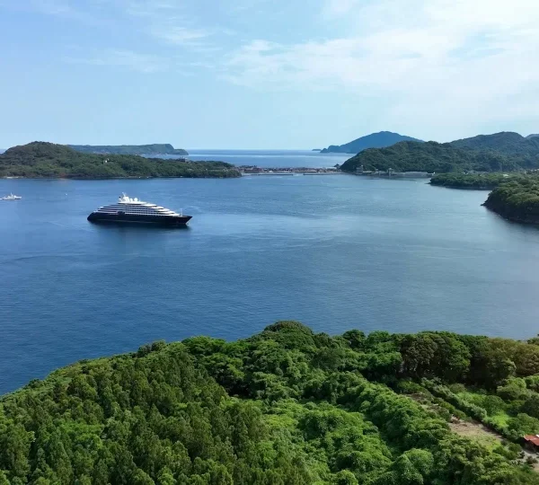 Aerial view of a blue bay with a large cruise ship near green islands; a pier extends from the right into the water.