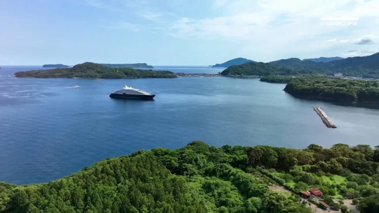 Aerial view of a blue bay with a large cruise ship near green islands; a pier extends from the right into the water.