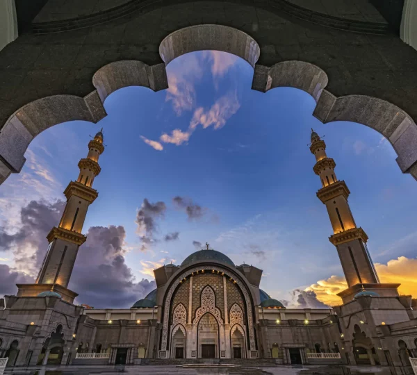 Ornate mosque entrance with tall minarets and a decorative archway at dusk in a spacious courtyard.