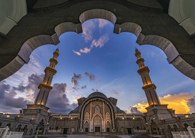 Ornate mosque entrance with tall minarets and a decorative archway at dusk in a spacious courtyard.