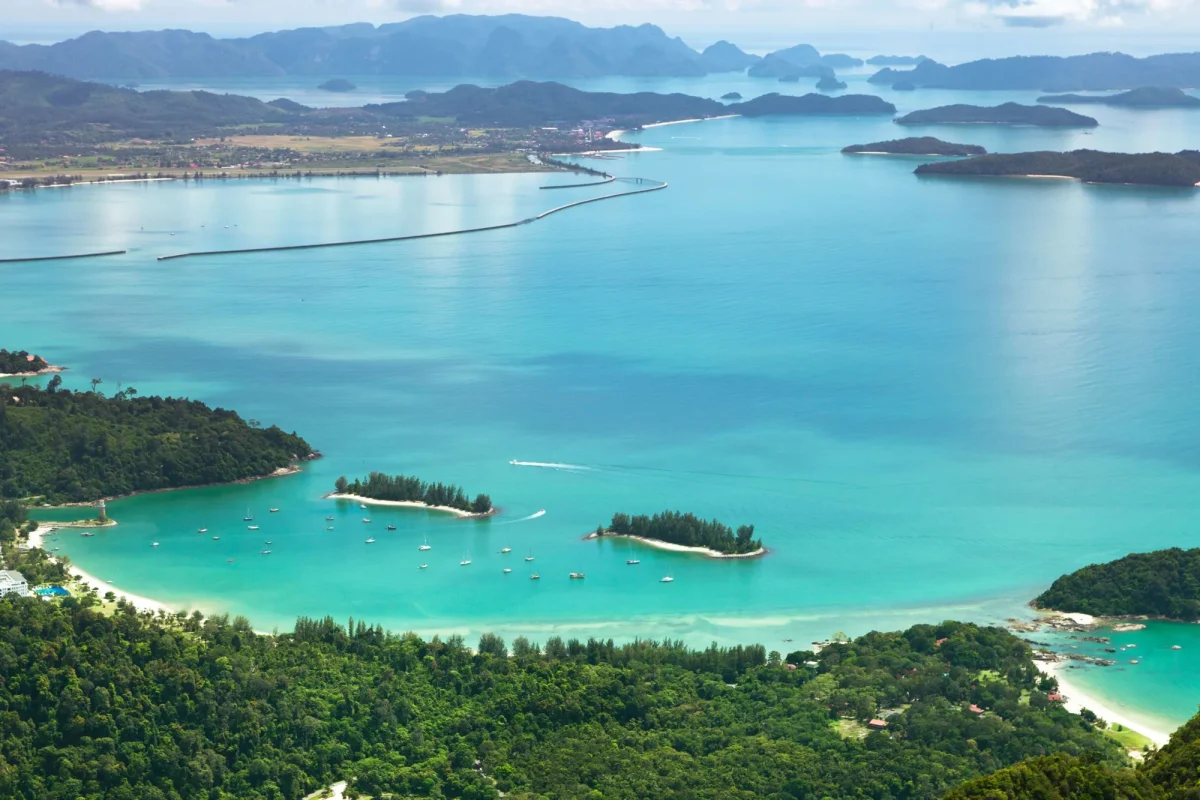 Aerial view of a tropical coastline with turquoise water, forested foreground, and scattered boats in a calm bay.
