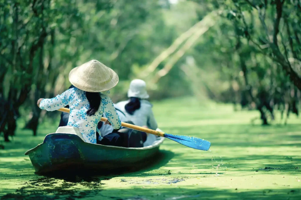 Woman in a conical hat rows a wooden boat through a green waterway lined with trees.