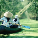 Woman in a conical hat rows a wooden boat through a green waterway lined with trees.
