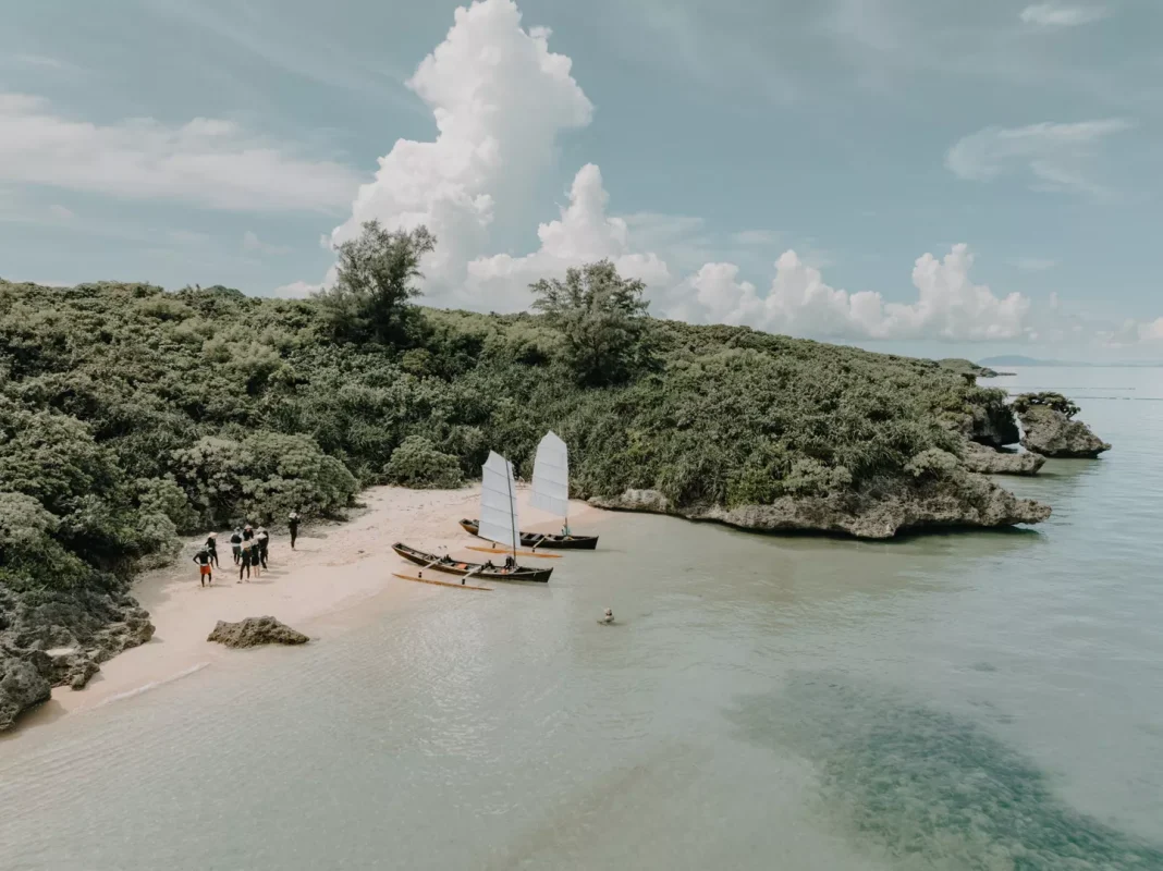 Tropical beach with a forested backdrop and small sailboats pulled onto the sandy shore; a group of people nearby by the water.