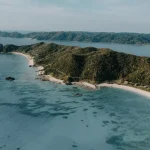 Aerial view of a forested peninsula stretching into a calm blue bay, with white-sand beaches along the curved shoreline and rocky outcrops.