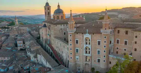 Sunset view of a stone fortress with towers and arched windows atop a hillside town.