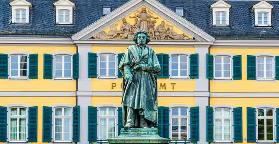 Bronze statue of a man in historical clothing stands before a yellow building with green shutters.