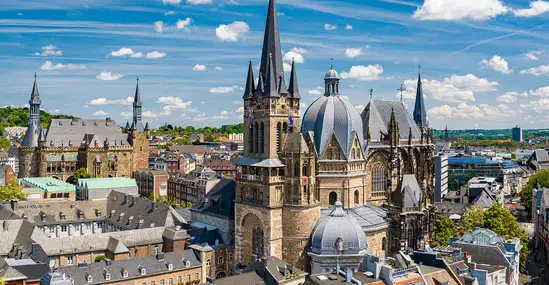 Aerial view of a historic European city with a Gothic cathedral and multiple spires rising above red-tiled rooftops under a blue sky.