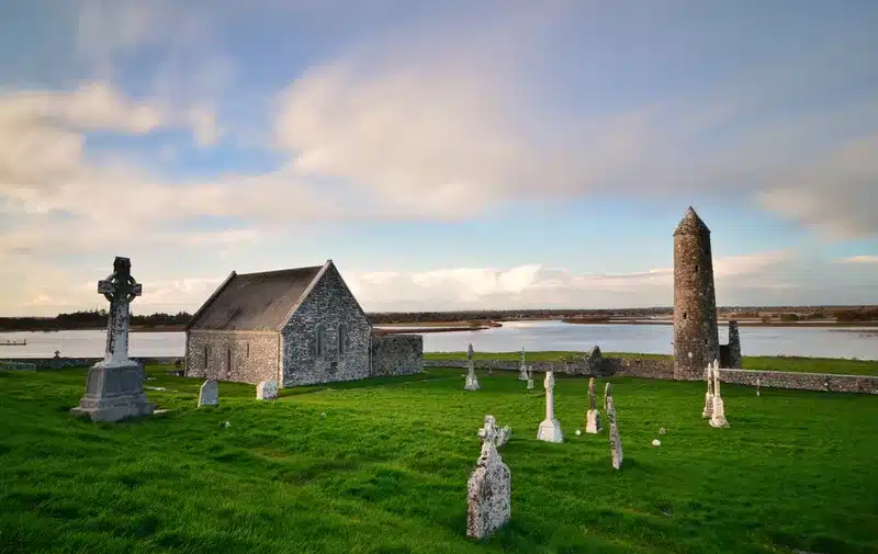 Stone church with a tall round tower, set among a graveyard on a grassy, coastal field overlooking the water.