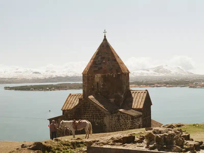 Stone church with a steep roof and cross on top overlooking a lake and snow-capped mountains in the distance.