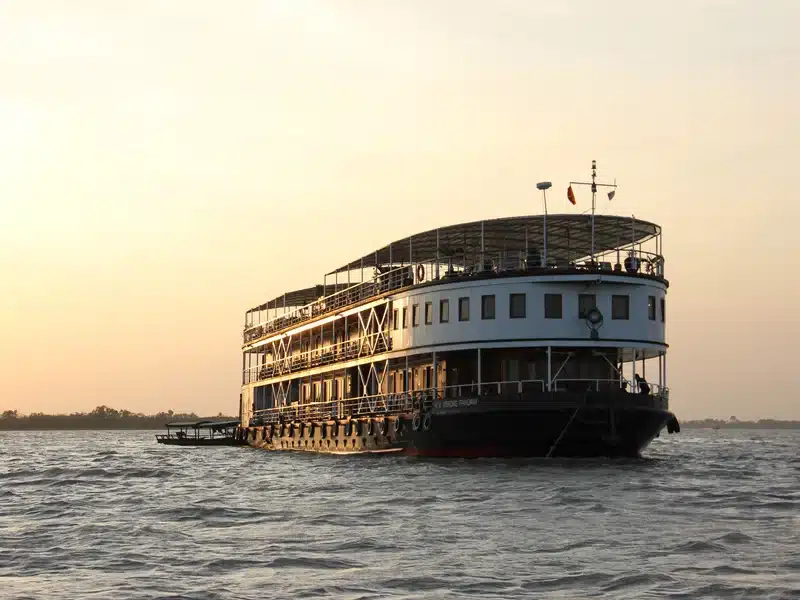 Sunset-lit two-level passenger steamboat cruising on calm river with open upper deck railings.