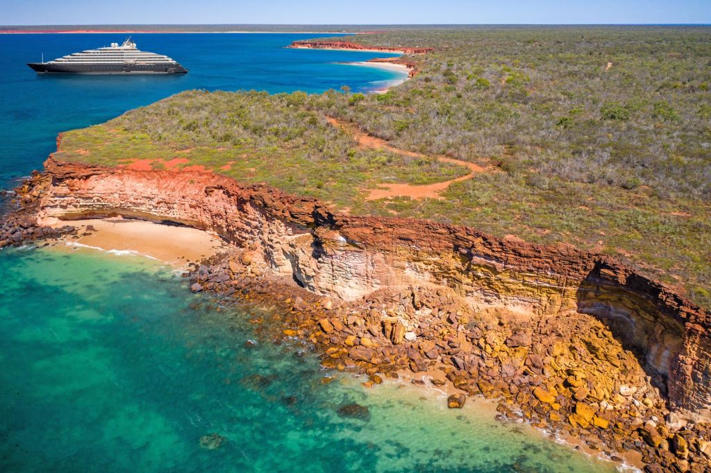 Aerial view of a large cruise ship near a red cliff coastline with turquoise water and sparse coastal vegetation.