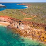 Aerial view of a large cruise ship near a red cliff coastline with turquoise water and sparse coastal vegetation.