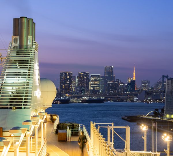 Cruise ship deck at dusk with city skyline across the water and illuminated railing in the foreground.