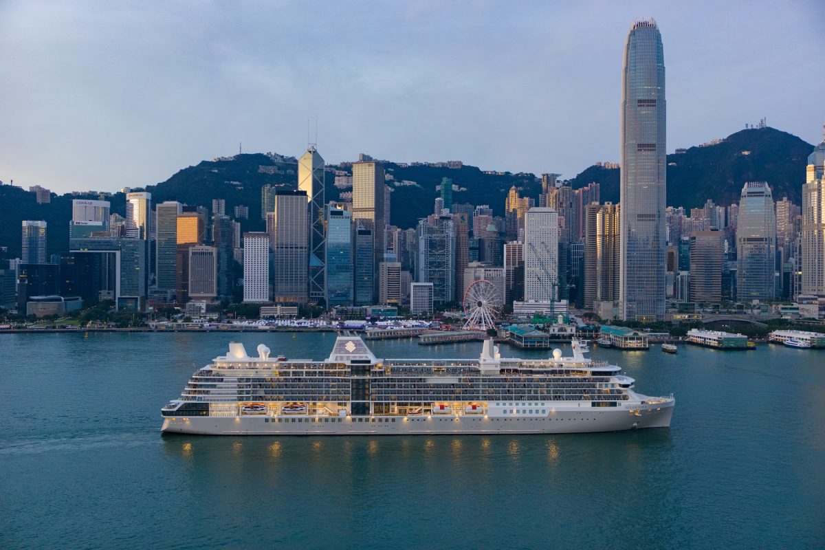 White cruise ship moored in Victoria Harbour with Hong Kong’s skyline and green hills behind it.