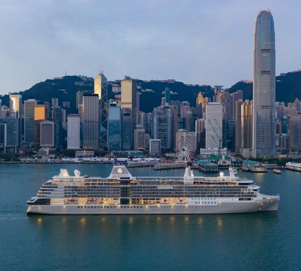 White cruise ship moored in Victoria Harbour with Hong Kong’s skyline and green hills behind it.