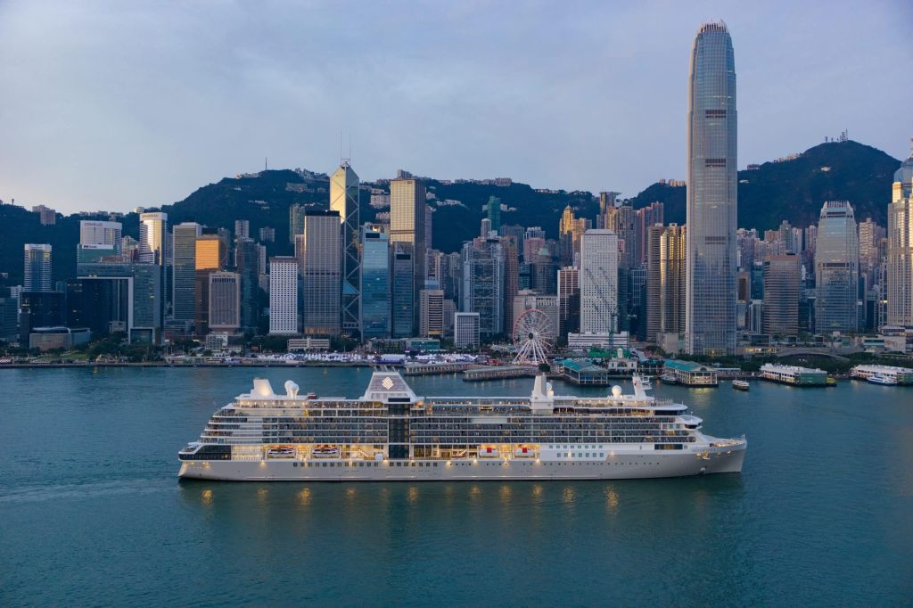 White cruise ship moored in Victoria Harbour with Hong Kong’s skyline and green hills behind it.