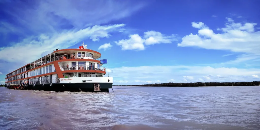 Large multi-deck river cruise ship on a muddy river under a bright blue sky with scattered clouds.