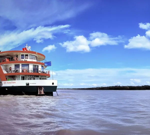 Large multi-deck river cruise ship on a muddy river under a bright blue sky with scattered clouds.