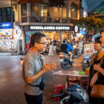 Guide chats with two women on a night street in front of a Highlands Coffee shop.