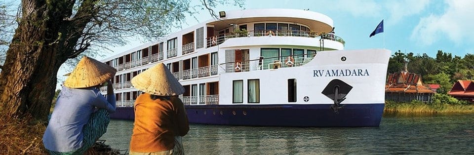 River cruise ship RV AMADARA on a calm river, with two people wearing conical hats sitting by the water at the shore.