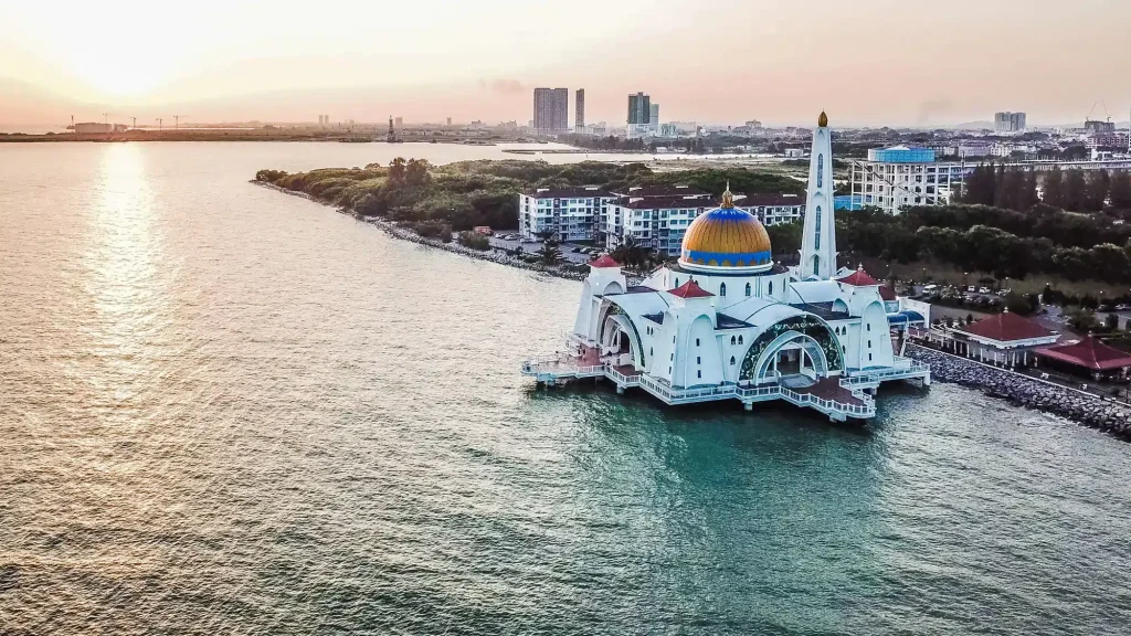Islamic-style mosque on a pier jutting into calm water at sunset, with city skyline in the distance.
