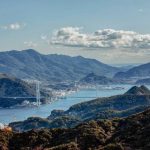 Coastal valley with a suspension bridge spanning a blue bay, surrounded by rolling hills and distant mountains under a partly cloudy sky.