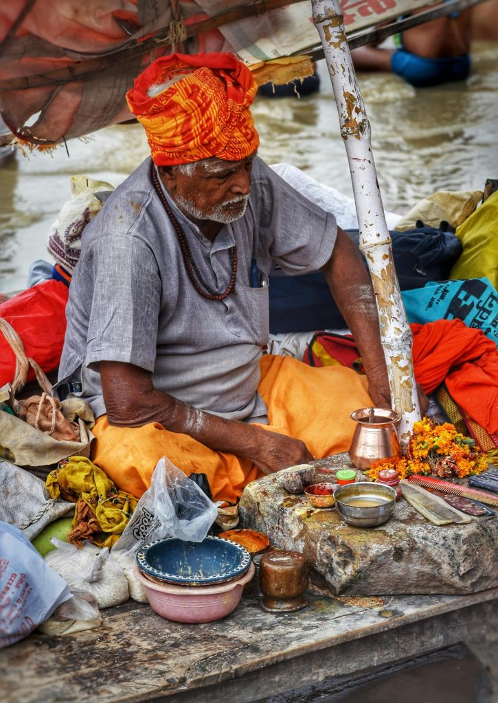 Elderly Indian man in an orange turban sits on a boat surrounded by colorful fabrics and metal bowls by the riverbank, performing a ritual.