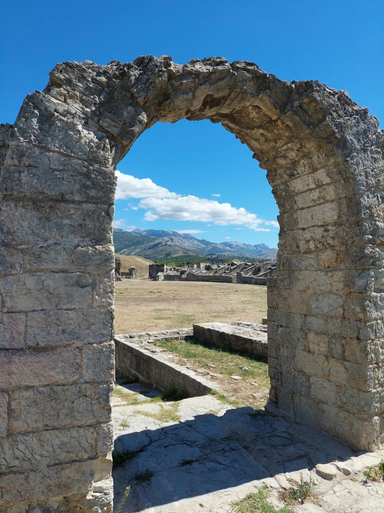 Ruined stone archway framing a landscape of mountains under a blue sky, sunny day.