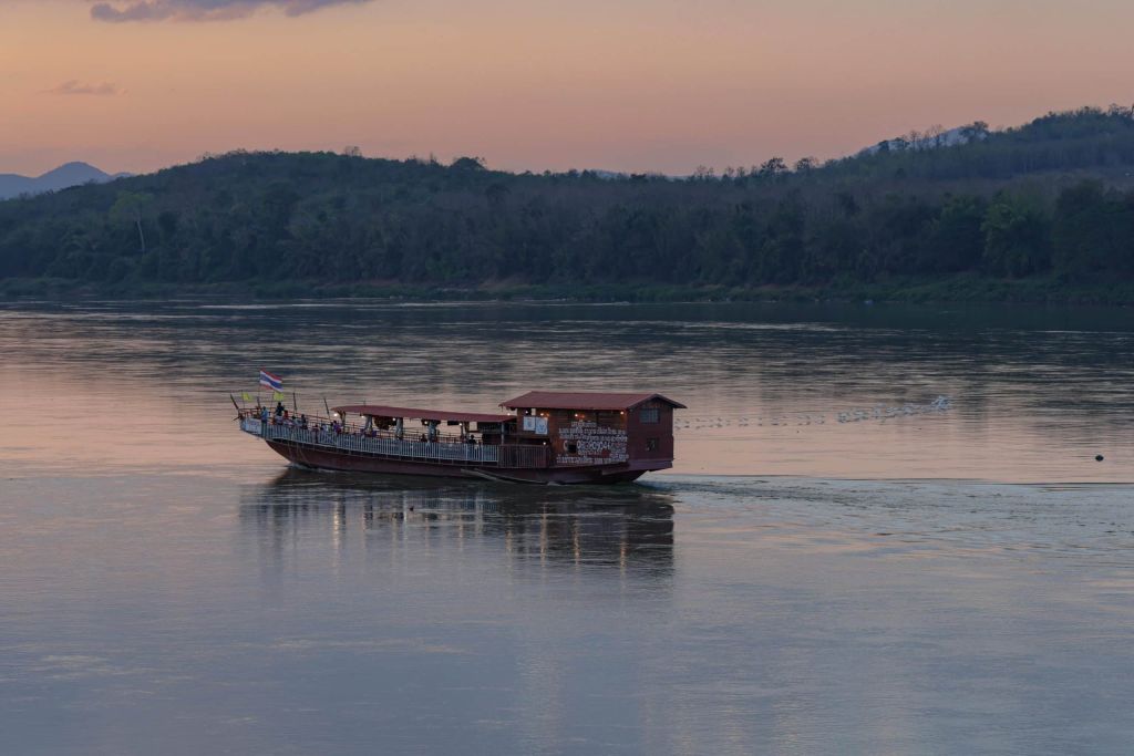 Long narrow covered river boat sailing on calm water at dusk.