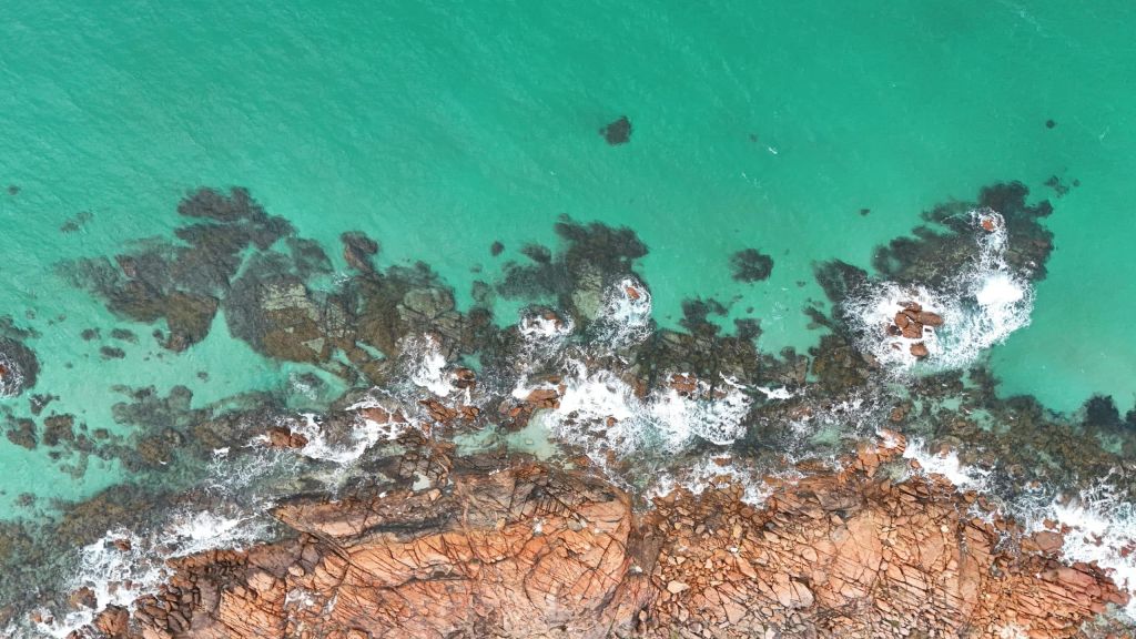 Aerial view of turquoise water washing over rugged brown rocks along a coastline with white foam nearby.