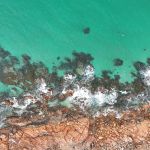 Aerial view of turquoise water washing over rugged brown rocks along a coastline with white foam nearby.