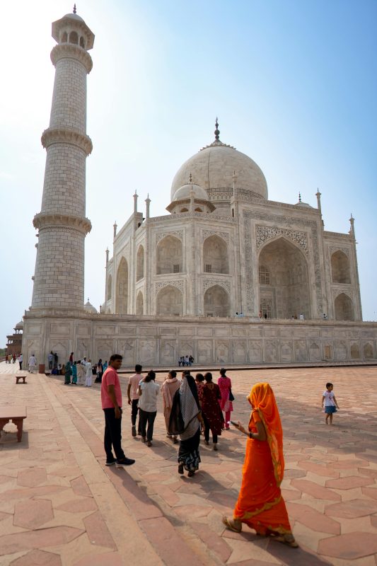 Taj Mahal with tall minaret on the left and visitors walking in the marble courtyard in the foreground.