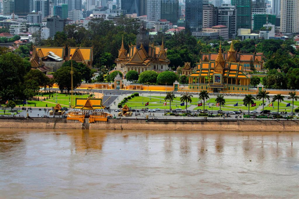 Ornate golden temple complex by a river with a modern city skyline behind it, green lawns and palm trees in the foreground.