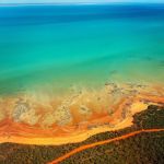 Aerial view of turquoise ocean meeting an orange sandy shore with a winding road through a dense green forest nearby.